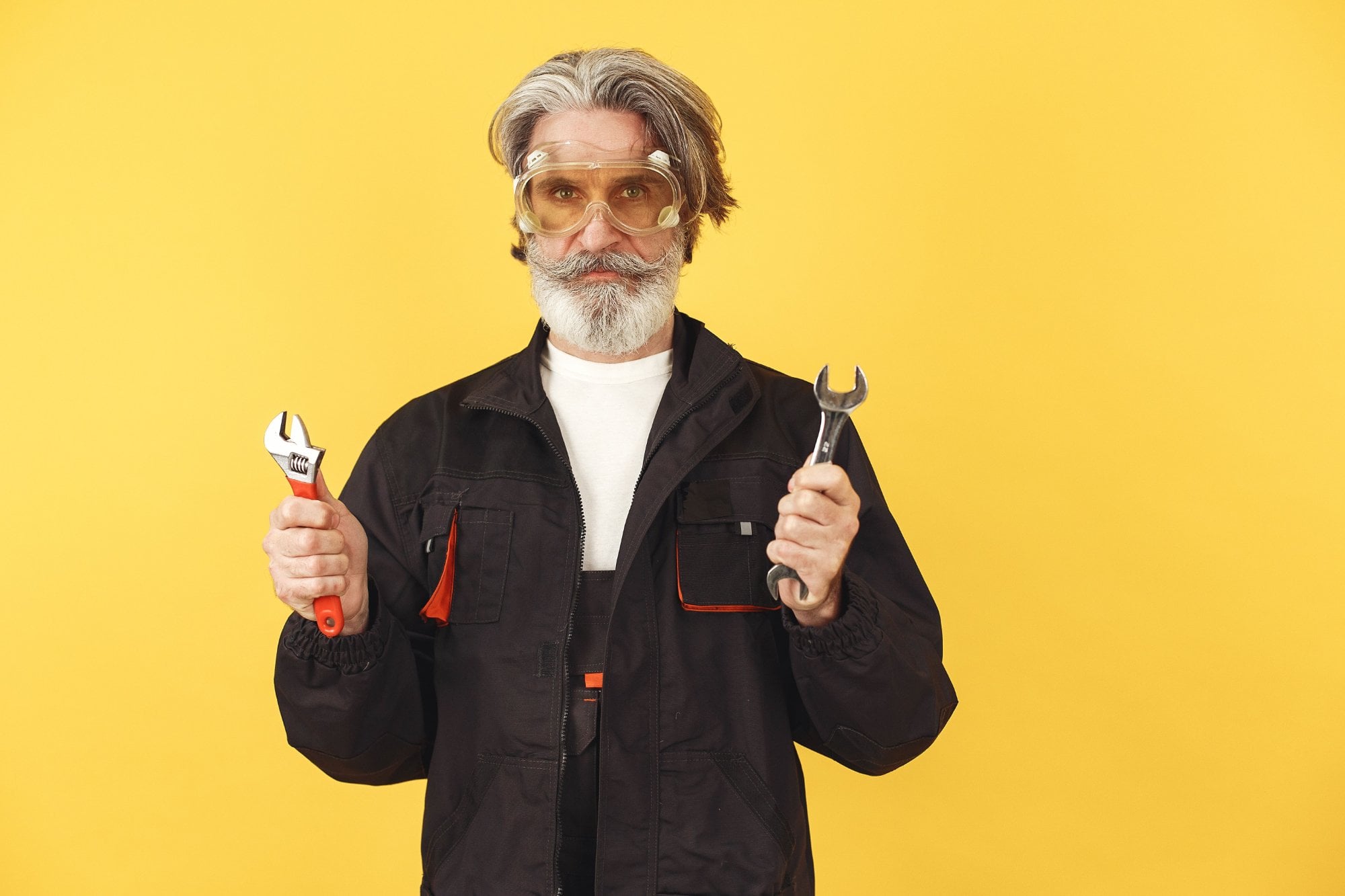 Hombre mayor con barba y gafas de seguridad sostiene una llave ajustable y una llave de boca